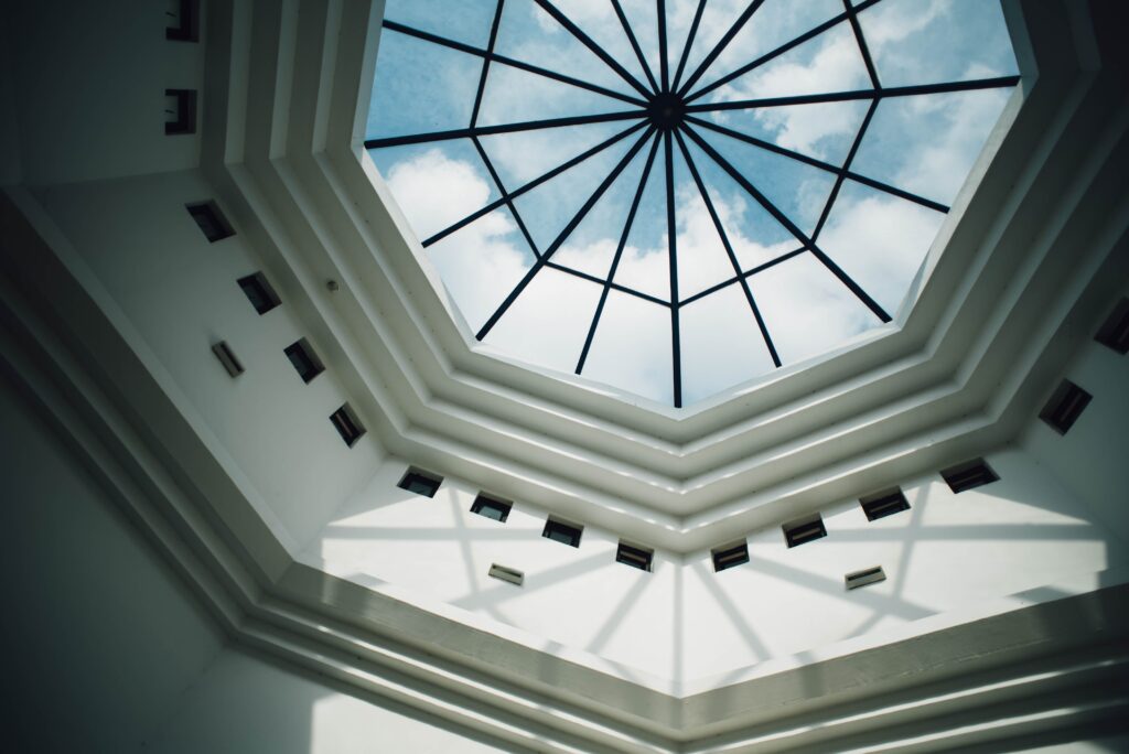 Octagonal skylight with black metal frame letting in natural light, viewed from below in a white interior, revealing a blue sky with clouds.