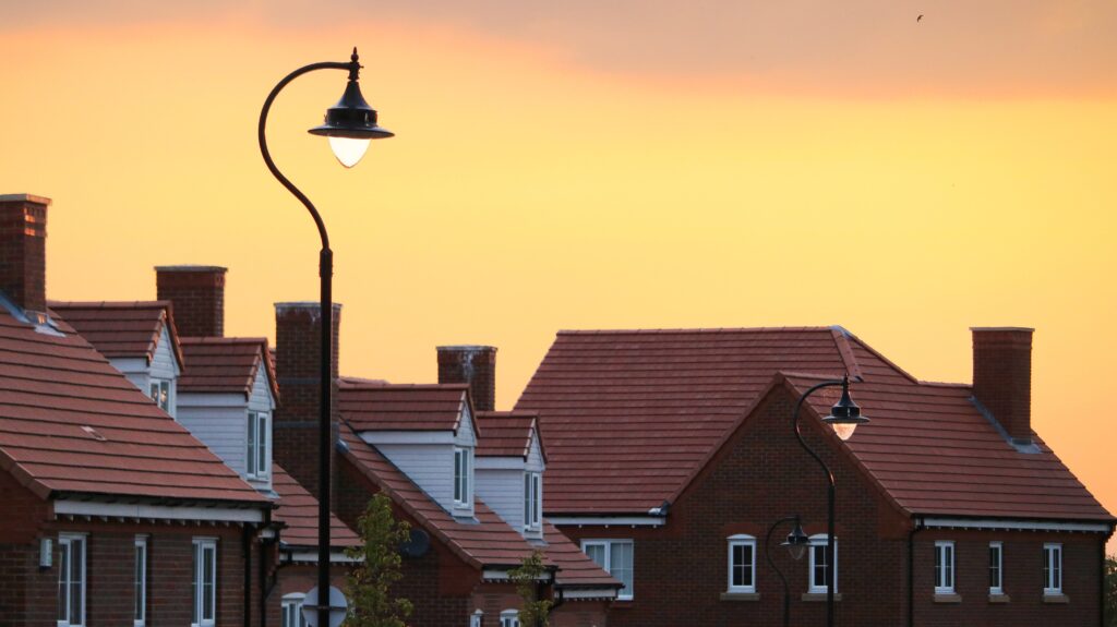 Street lamps and modern brick houses with red tiled roofs are silhouetted against a yellow-orange sunset sky.