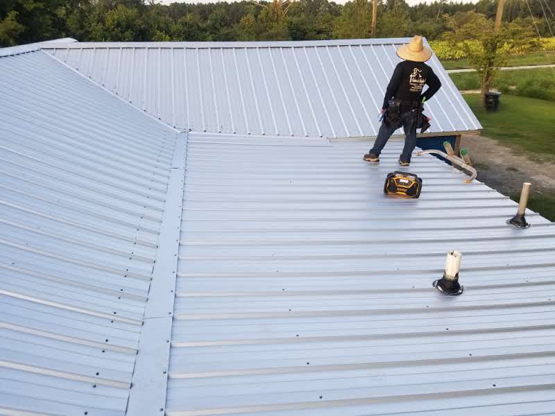 A worker in a large hat stands on a white metal roof holding a tool, facing away. Roofing materials and tools are nearby.