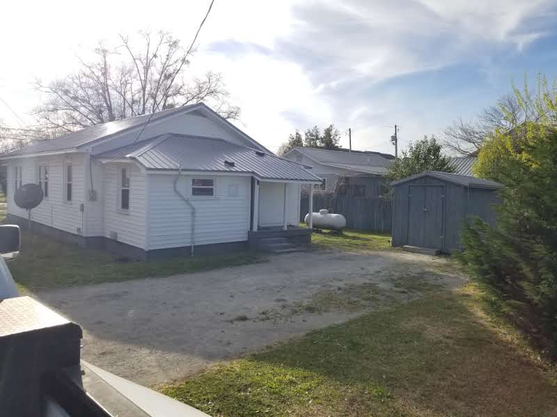 A small white house with a metal roof, a shed in the yard, and propane tanks near the back, under a partially cloudy sky.