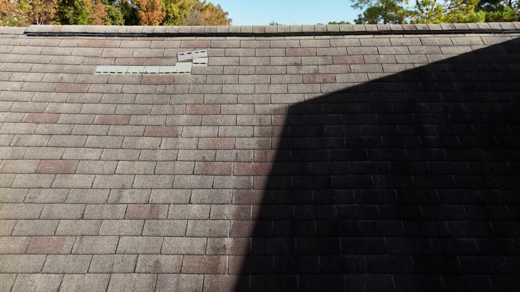 A shingled roof with a section of missing shingles and a large shadow cast by an adjacent structure. Trees are visible in the background.