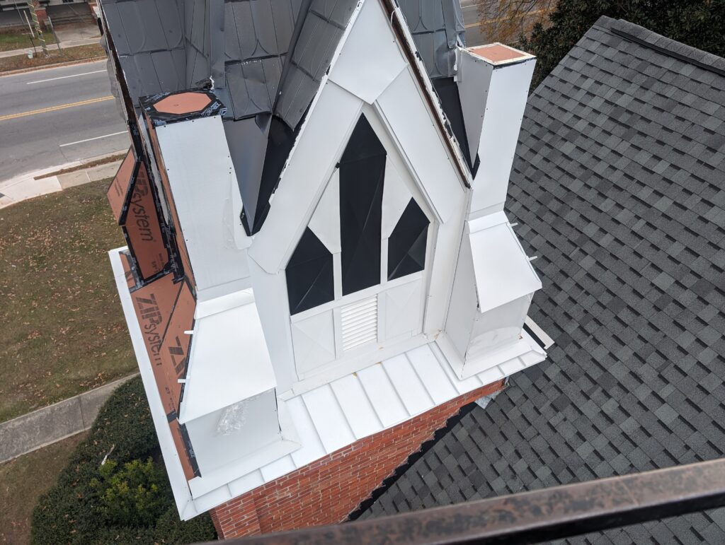 Aerial view of a church with a newly repaired roof and steeple showing white and black sections, adjacent to a road and green area.