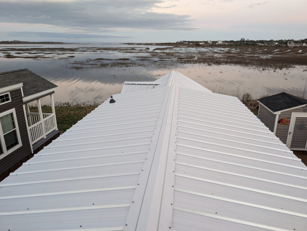 A white metal roof stretches towards a serene body of water with grassy marshlands and houses in the distance under a cloudy sky.