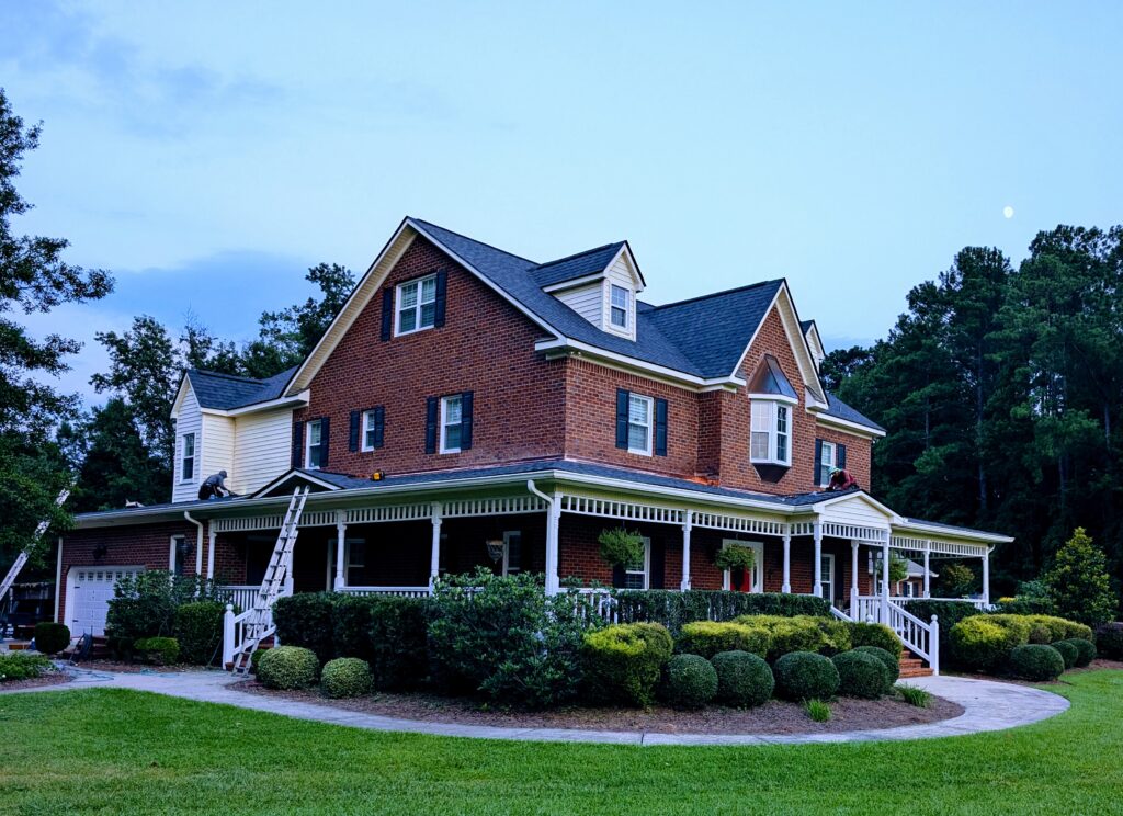 A large brick house with a wraparound porch, green lawn, and surrounding trees under a blue sky at dusk. Two ladders are propped up.