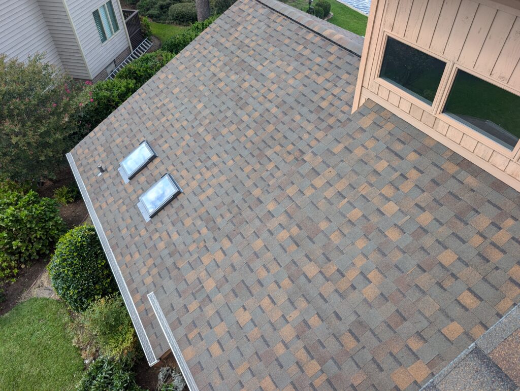 Aerial view of a residential roof with two skylights and brown asphalt shingles next to a wooden structure and greenery.