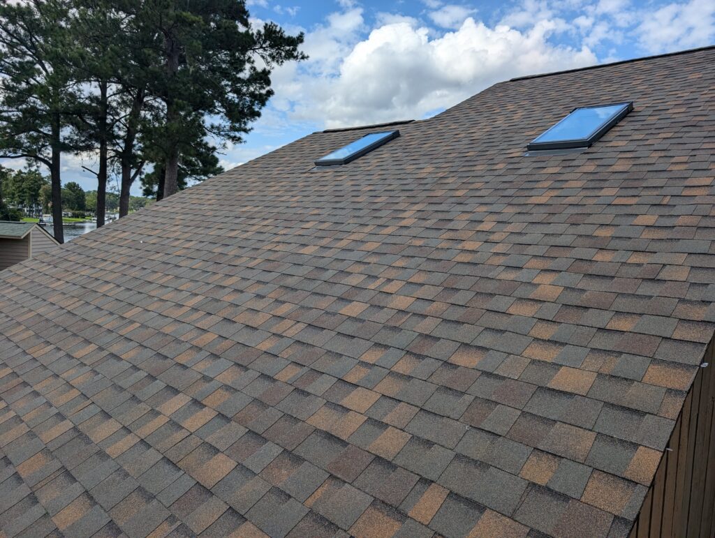 Shingled roof with two skylights, set against a backdrop of trees and a partly cloudy sky.