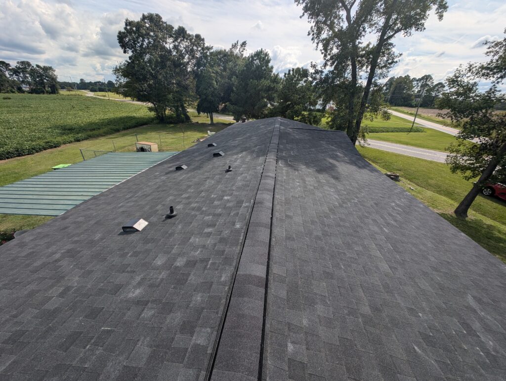 Aerial view of a shingled roof on a residential building surrounded by trees and fields with a road on the right side.